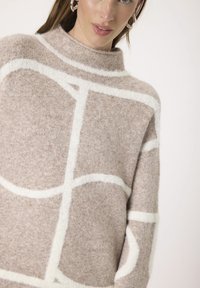Lysebrunt sweater med høj krave, prydet med hvide abstrakte mønstre. Lavet af blødt, tekstureret stof, har den dropped shoulders og en afslappet pasform.