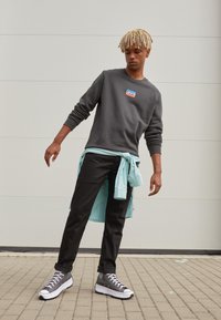 Sudadera gris oscura con el logo de Levi's, camisa azul clara atada a la cintura, pantalones negros y zapatillas altas grises con suelas blancas.