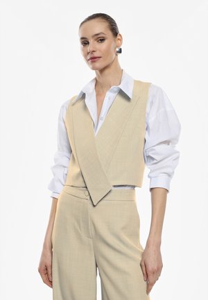 Gilet beige su misura con design a scollo a V sopra una camicia bianca. La texture è liscia, con revers larghi e una silhouette aderente.
