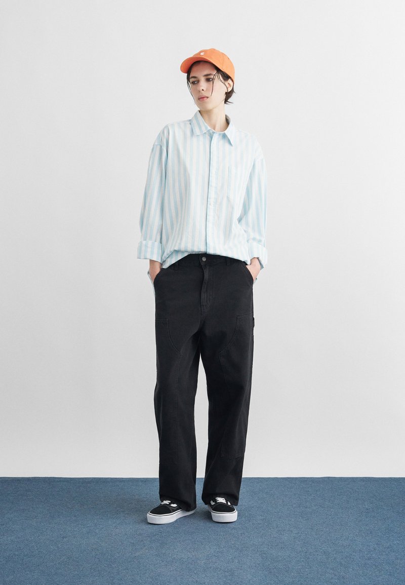 Giovane con cappellino arancione, camicia a righe azzurre, pantaloni neri larghi e sneaker nere, in piedi su un tappeto blu davanti a una parete bianca liscia.