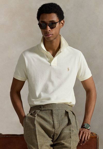 Πόλο μπλούζα σε ανοιχτό κίτρινο χρώμα με γιακά δύο κουμπιών και κεντημένο λογότυπο. Συνδυάζεται με καρό καφέ παντελόνια και στρογγυλά γυαλιά ηλίου.