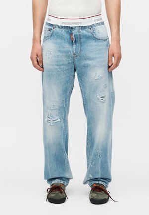 Άνδρας που φορά ανοιχτό μπλε εξαιρετικά φθαρμένο τζιν με φαρδιά εφαρμογή και γυρισμένα ρεβέρ, μαύρα και πορτοκαλί αθλητικά παπούτσια, και ορατή λευκή μέση εσώρουχου DSQUARED2.