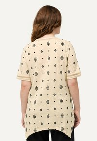 Beige-neulepaita, jossa on mustia geometrisia kuvioita, lyhyet hihat ja kaareva helma. Kankaassa on sileä rakenne ja rento istuvuus.