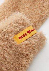 Imagem em close de luvas fofas de cor castanha clara com um rótulo amarelo cosido que apresenta texto em vermelho e um logótipo de coração.