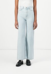 Jeans de tiro alto en un azul claro, de pierna ancha, fabricados en denim, con un diseño clásico de cinco bolsillos y cierre de botón. Mocasines negros completan el look.