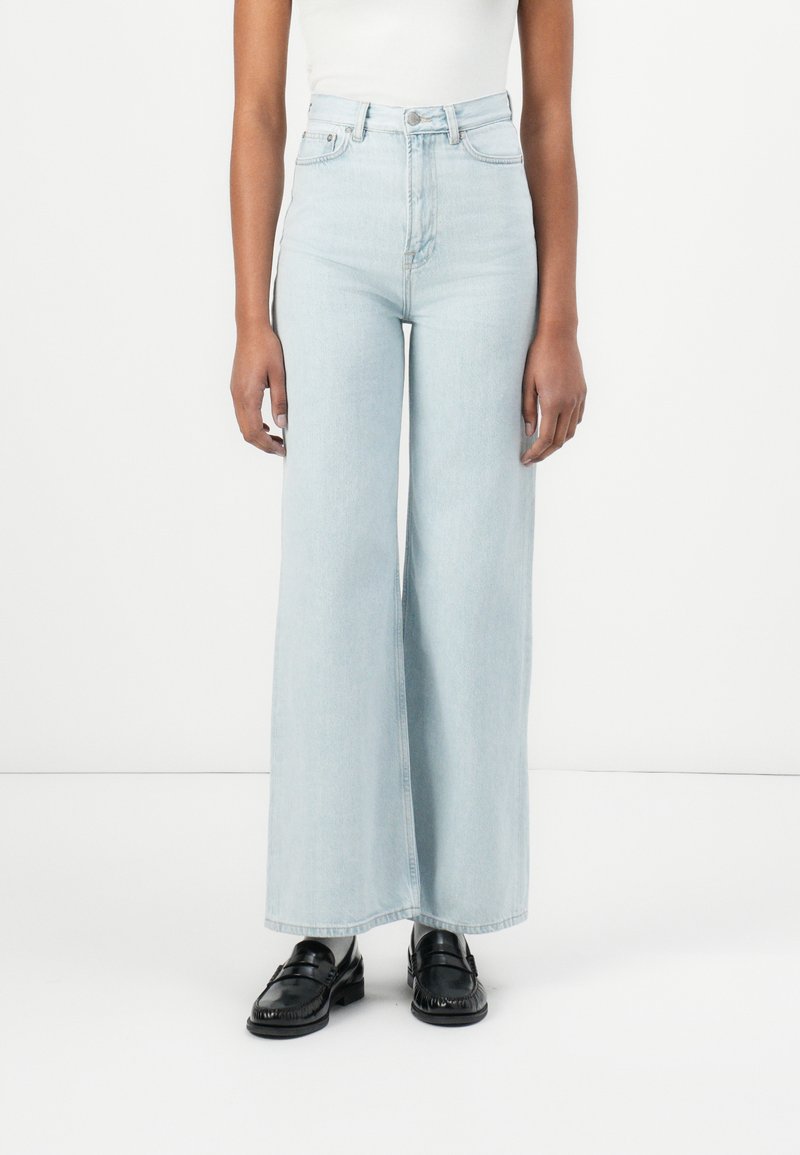 Jeans de tiro alto en un azul claro, de pierna ancha, fabricados en denim, con un diseño clásico de cinco bolsillos y cierre de botón. Mocasines negros completan el look.