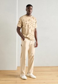 Beige T-Shirt mit kurzen Ärmeln und geometrischen Mustern, kombiniert mit hellbeigen Cargo-Hosen und weißen Sneakers mit grauen Akzenten.