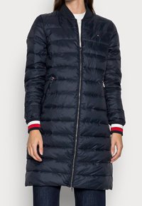 Granatowa puchowa kurtka z zamkiem z przodu, żebrowanym kołnierzem i paskowanymi mankietami. Posiada poziomy wzór pikowania oraz boczne kieszenie.