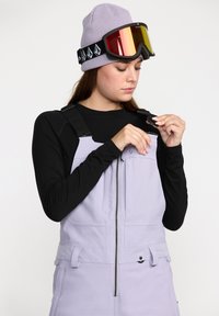 Salopette de ski lilas avec bretelles noires réglables, fermeture éclair à l'avant et poches, assortie à un bonnet en tricot lavande et à des lunettes de ski miroir.
