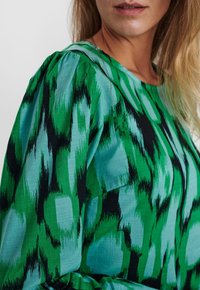 Blouse verte avec des motifs abstraits dans des nuances de vert et de noir. Fabriquée à partir d'un tissu doux, elle possède un col rond et une coupe ample.