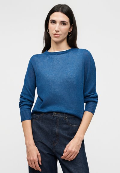 Femme aux longs cheveux noirs portant des boucles d'oreilles en or en forme de cercle, un pull en tricot bleu à manches longues et un jean bleu foncé, devant un fond uni.