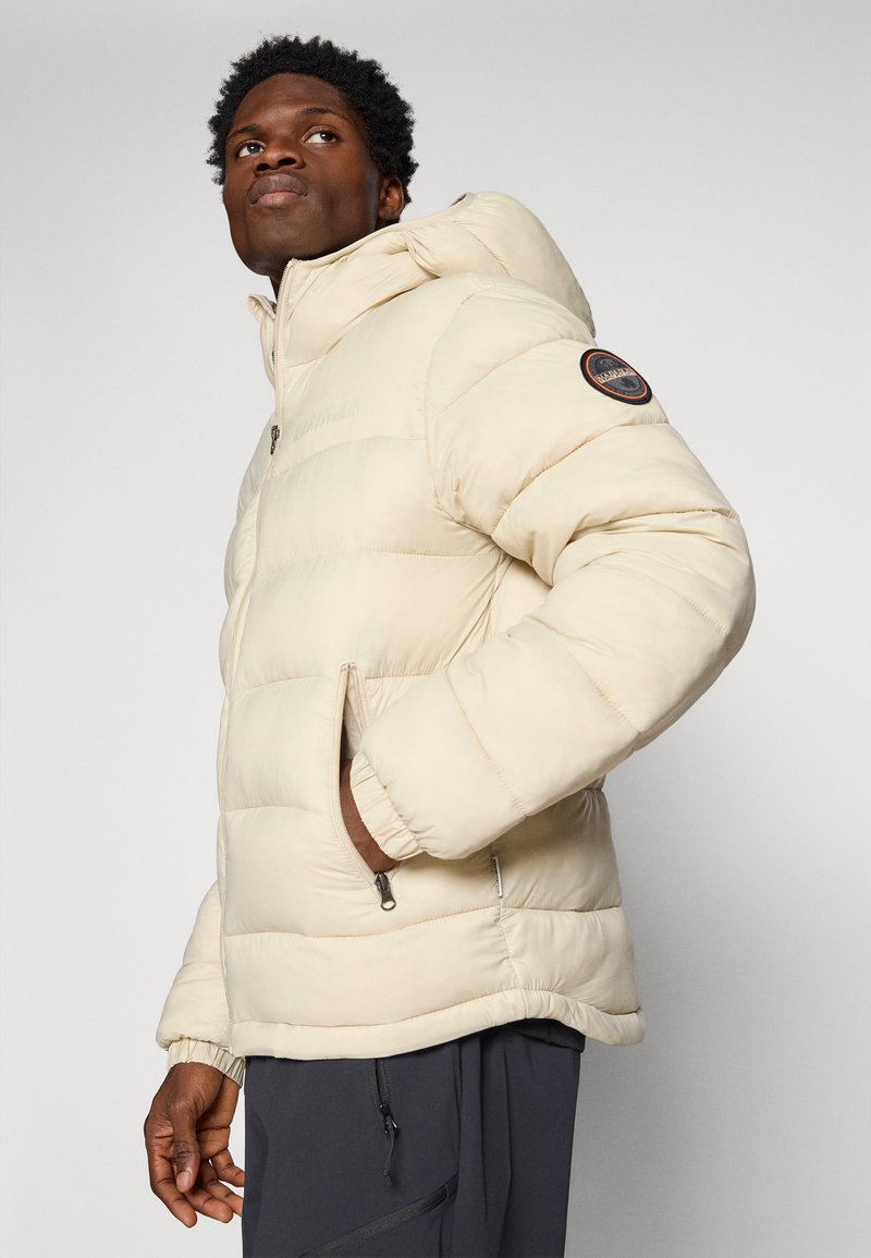 Jasno beżowa kurtka puchowa z kapturem, z poziomym pikowaniem, bocznymi kieszeniami i okrągłą łatą z logo na lewym rękawie.