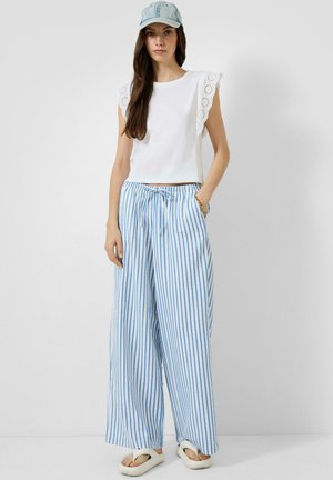 Vrouw draagt een witte mouwloze top met kanten randen, blauw-wit gestreepte broeken met wijde pijpen, een spijkerpet en witte sandalen.