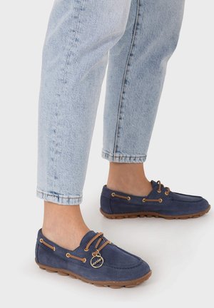 Persona que lleva vaqueros azul claro remangados y mocasines de ante azul marino con suelas de goma y un adorno dorado en los cordones.