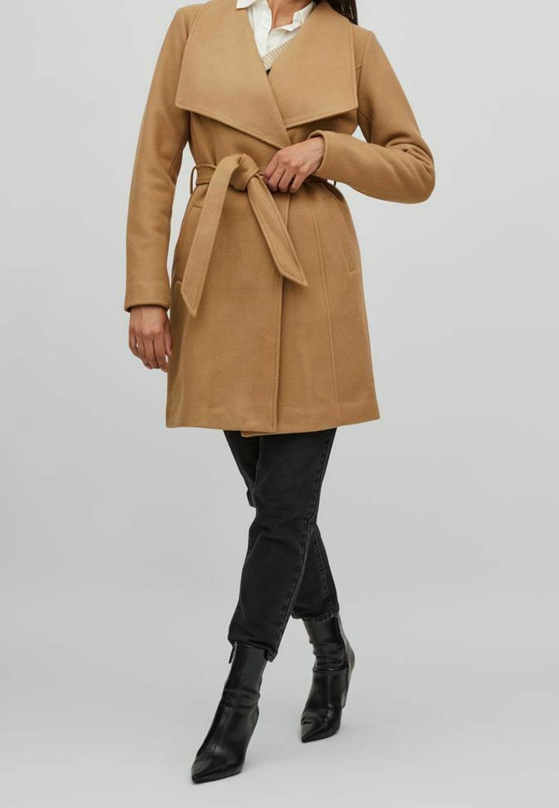 Femme ajustant une ceinture nouée sur un manteau long couleur chameau, portant un pantalon noir et des bottines noires sur un fond gris clair.