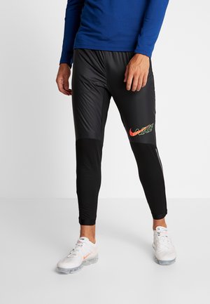 Mężczyzna ubrany w czarne sportowe spodnie Nike z kolorowym logo, niebieską bluzę z długim rękawem i białe sneakersy Nike, stojący na białej podłodze.