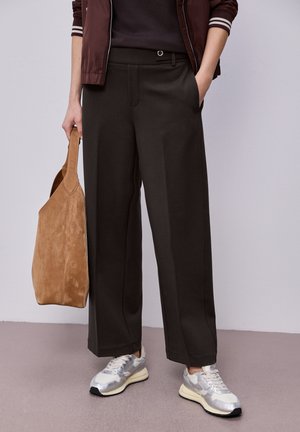 Personne vêtue d'un pantalon large et sombre, de baskets argentées, d'une veste marron, tenant un grand sac fourre-tout en daim beige à ses côtés.