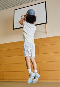 Person in a white sleeveless shirt and white shorts with blue accents jumps to shoot a blue basketball into a hoop against a wooden wall.