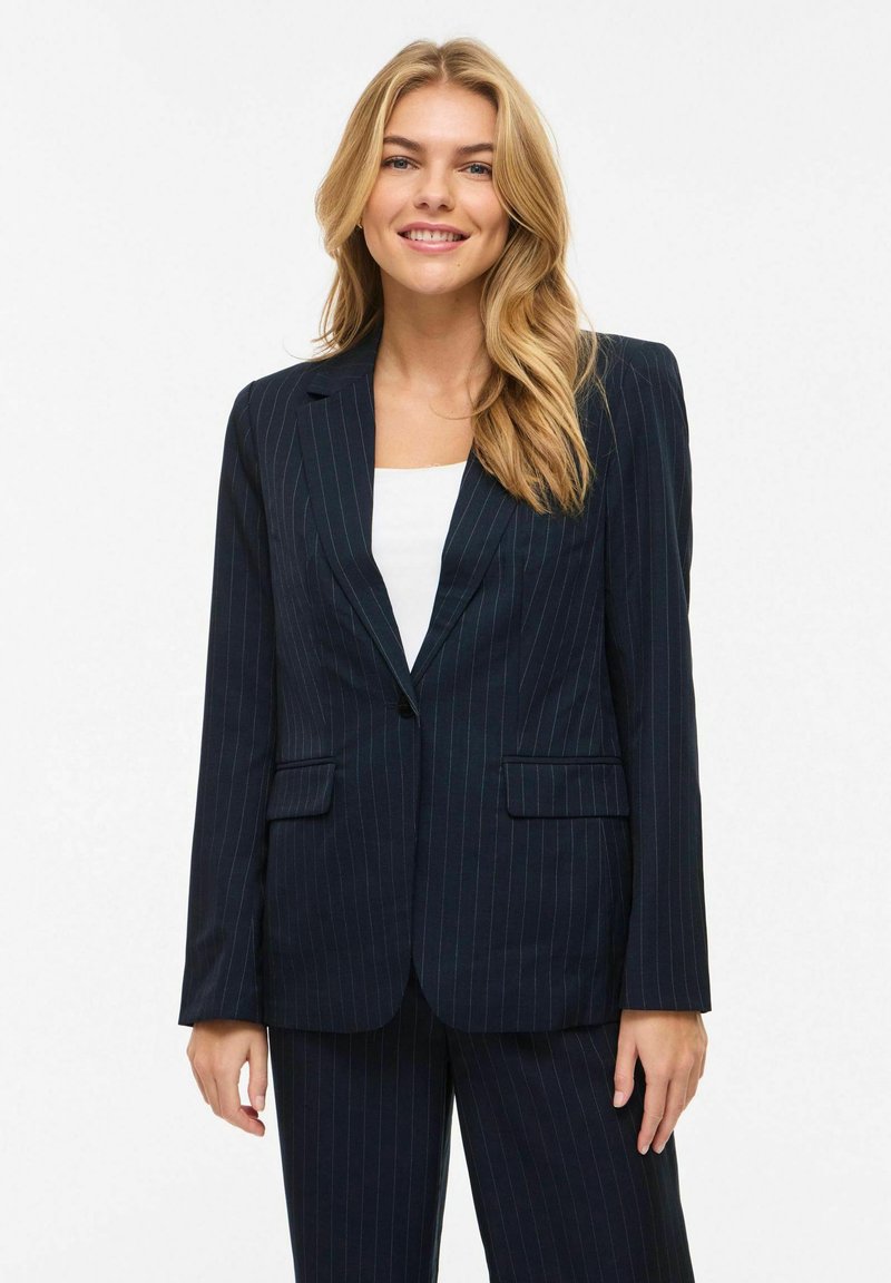 Femme blonde souriante, portant un blazer bleu marine à fines rayures et un pantalon assorti avec un haut blanc en dessous, debout devant un fond uni.