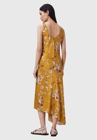 Abito floreale in giallo senape con schiena a V. Presenta un orlo asimmetrico fluido e una texture in satin. Sandali argentati completano l'outfit.