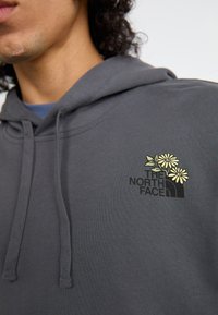 Γκρίζα φούτερ φτιαγμένη από μαλακό ύφασμα, με μπροστινό λογότυπο που περιέχει πράσινα φύλλα και κίτρινα λουλούδια. Διαθέτει κουκούλα με κορδόνι και ραφές ribbed.