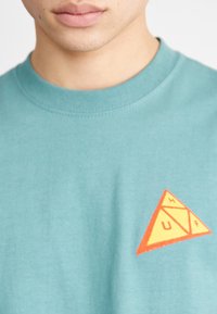 Camiseta de algodón turquesa con cuello redondo, que presenta un logo triangular naranja y amarillo en el lado izquierdo del pecho. Textura suave, diseño casual.