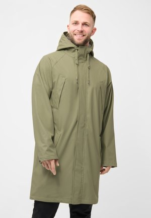 Mann trägt einen langen olivgrünen Kapuzenmantel mit Fronttaschen, steht und lächelt vor einem einfarbigen hellen Hintergrund.