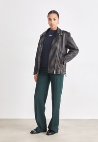 Giubbotto bomber in pelle nera con zip argentate, indossato sopra un maglione blu navy, abbinato a pantaloni larghi verdi e scarpe nere basse.
