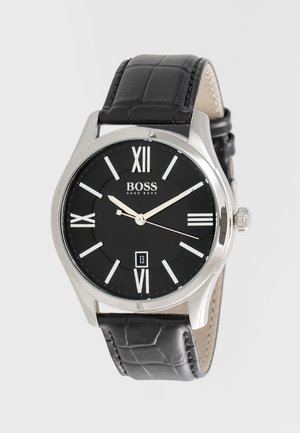 Montre-bracelet ronde noire Hugo Boss avec boîtier argenté, chiffres romains, guichet date à 6 heures et bracelet en cuir noir texturé.