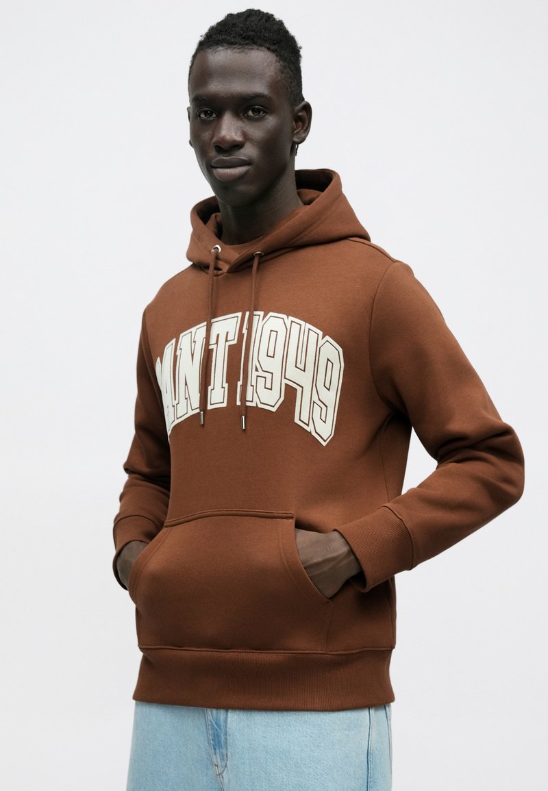 Brown hoodie with a front kangaroo pocket, featuring a large white graphic "MNT 1949" across the chest. Soft fabric with ribbed cuffs.