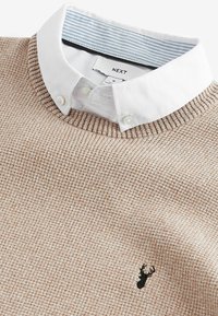 Strikbeige sweater med et struktureret mønster og ribbet krave. Har en hvid skjorte-krave under. Sort logo-udtryk på brystet.