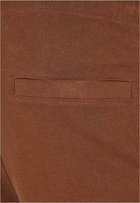 Bruine katoenen stof met een gladde textuur, voorzien van een achterzak met een horizontale klep en versterkte stiksels langs de randen.