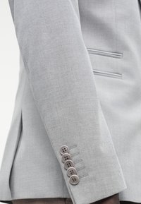 Manica di blazer grigio chiaro con tessuto testurizzato, dettaglio di cucitura sottile e cinque bottoni marroni sul polsino.