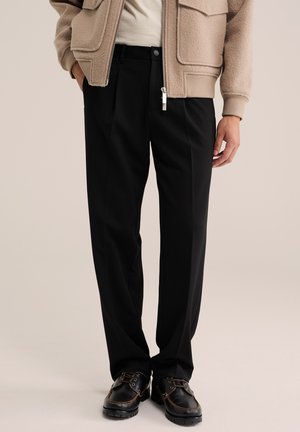 Personne portant un pantalon noir droit, des chaussures en cuir noir à lacets, une veste beige avec de grandes poches et une chemise blanche.
