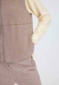 Beige Fleece-Weste mit abgerundeten Kanten, mit einem Reißverschluss und seitlichen Taschen, über einem weichen cremefarbenen Langarmshirt getragen.