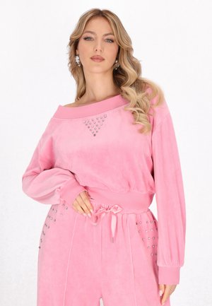Kobieta o długich blond włosach nosi różowy aksamitny top z odkrytymi ramionami oraz dopasowane spodnie ozdobione małymi srebrnymi ćwiekami.