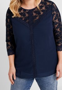 Blouse bleu foncé avec des manches trois-quarts en dentelle et un motif floral. Corps lisse avec un détail de dentelle le long du centre.