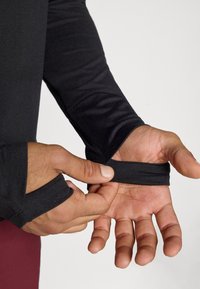 Melnā garenā piedurkņu krekls ar pirkstu caurumiem aprocēs. Tam ir maiga tekstūra un pieguļoša forma, kas ir piemērota aktīvai valkāšanai.