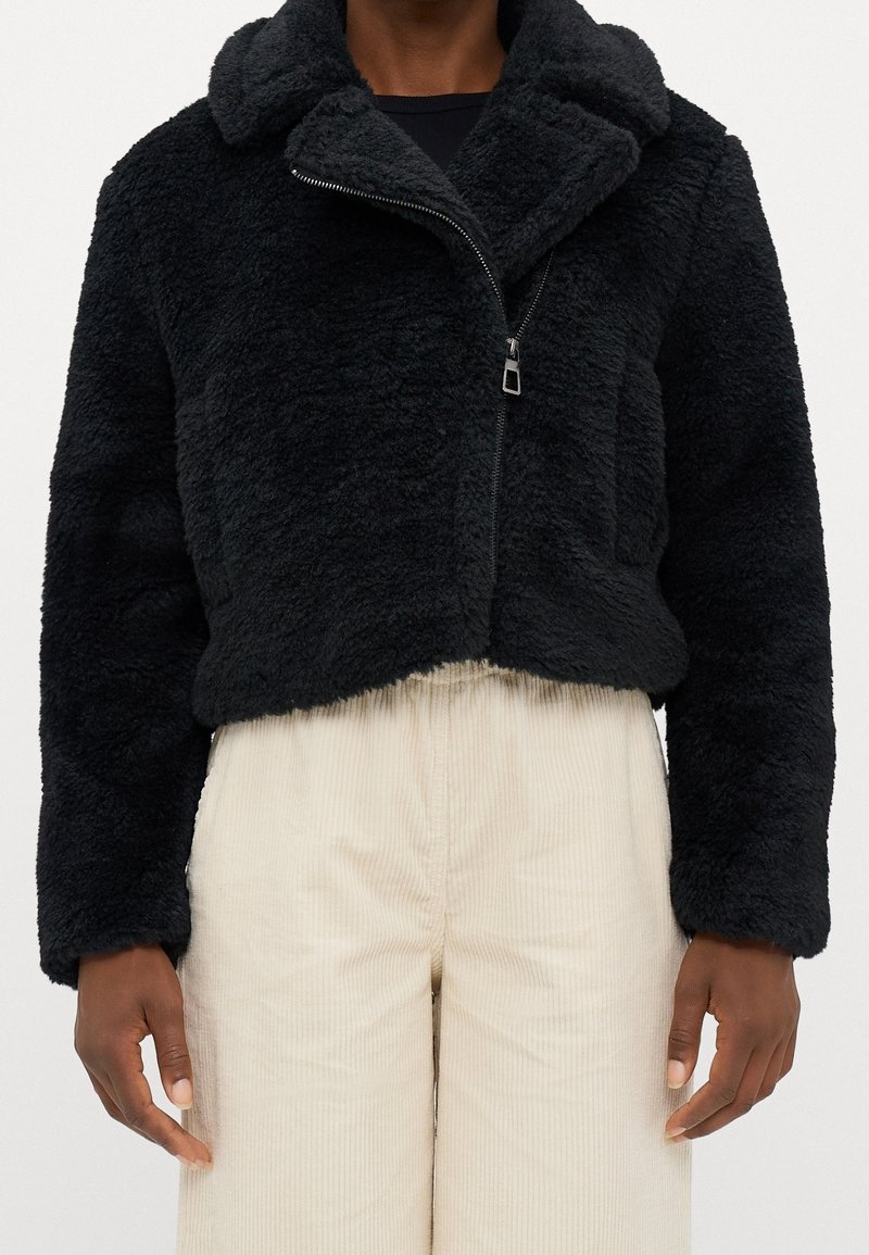 Schwarze, kurz geschnittene Jacke aus plüschigem, strukturiertem Stoff mit einem Reißverschluss und breitem Kragen. Getragen über einer hellen Cordhose.
