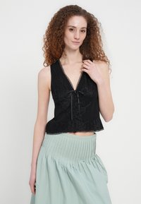 Γυναίκα με σγουρά κόκκινα μαλλιά, που φοράει μαύρο δαντελένιο τοπ χωρίς μανίκια και ανοιχτοπράσινη πλισέ φούστα, ποζάροντας μπροστά σε απλό λευκό φόντο.