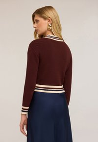 Maglione a maglia bordeaux con design cropped, caratterizzato da un orlo e polsini a coste con righe crema. Indossato sopra un indumento in satin blu scuro.