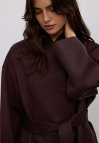 Tmavě hnědý oversized župan s páskem a širokými rukávy vyrobený z měkké tkaniny, s jednoduchým, minimalistickým designem a hladkou strukturou.