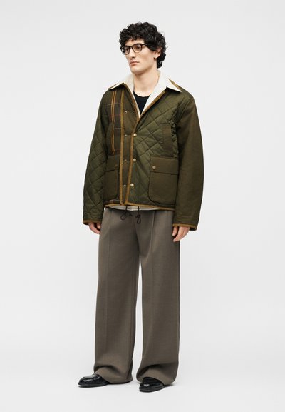 Νέος άνδρας με σγουρά μαλλιά και γυαλιά, φοράει πράσινο καπιτονέ μπουφάν, μπεζ υπερμεγέθη παντελόνια και μαύρα παπούτσια, στέκεται σε απλό φόντο.