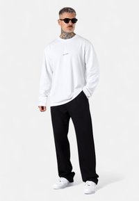 Weißes Langarmshirt mit kleinem Textlogo auf der Brust, kombiniert mit schwarzen Hosen und weißen Sneakers. Schicke Sonnenbrillen runden das Gesamtbild ab.