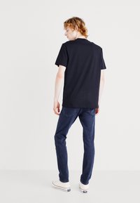 T-shirt en coton noir présenté de dos, associé à un jean foncé et des baskets blanches, sur fond blanc uni.