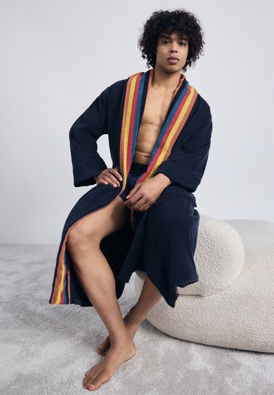 Jeune homme aux cheveux bouclés portant une robe de marine avec des rayures multicolores, assis pieds nus sur un siège avec un rembourrage texturé blanc.