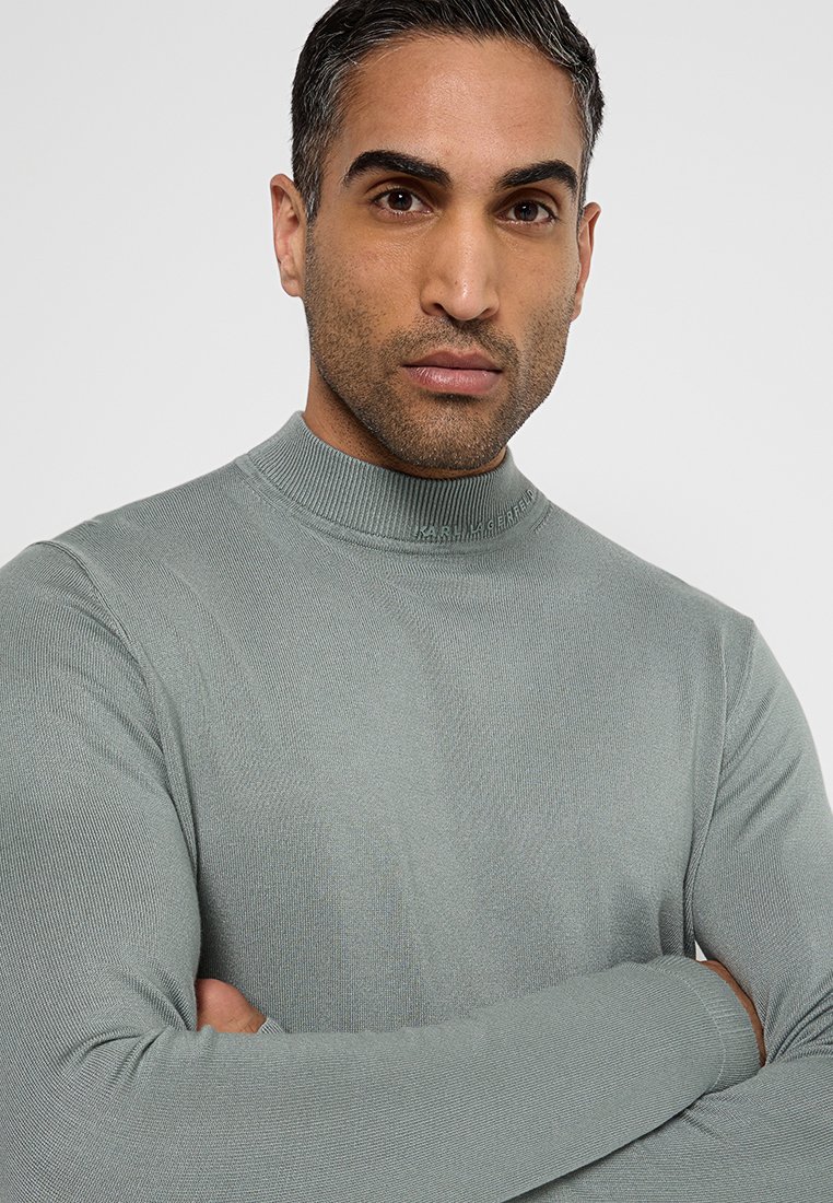 Ein Mann mit verschränkten Armen, der einen hellgrauen Strickpullover mit Stehkragen trägt, vor einem schlichten, hellen Hintergrund.