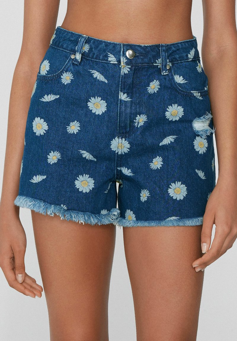 Szorty dżinsowe z postrzępionym wykończeniem, w niebieskim kolorze, pokryte rozrzuconymi białymi wzorami stokrotek. Pięć kieszeni, metalowy guzik.