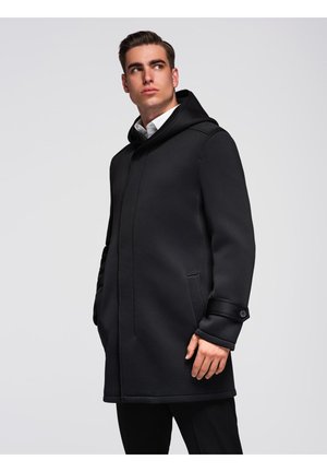 Jeune homme portant un manteau noir à capuche sur une chemise blanche, se tenant avec les mains dans les poches, regardant sur le côté sur un fond clair.