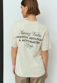 Tricou din bumbac crem, cu mâneci scurte și guler rotund. Pe spate se află un text negru care spune "Suivez Votre Ambition Artistique à Montmartre."
